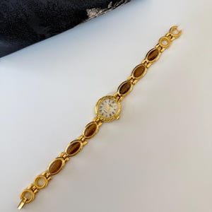 May include: A gold-toned wristwatch with a round face and Roman numeral hour markers. The watch band features oval-shaped brown stones set in gold links. The watch is displayed on a white surface.