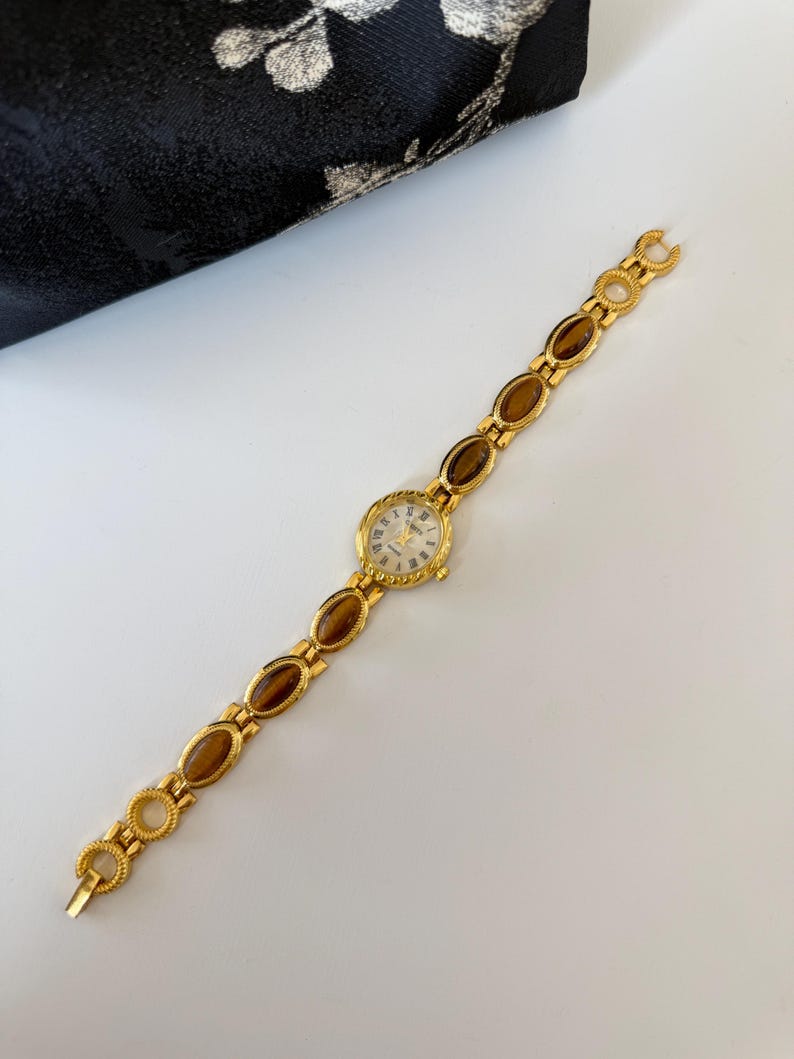 May include: A gold-toned wristwatch with a round face and Roman numeral hour markers. The watch band features oval-shaped brown stones set in gold links. The watch is displayed on a white surface.