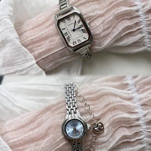 K&ouml;nnte beinhalten: Zwei silberne Armbanduhren mit Metallb&auml;ndern. Die obere Uhr hat ein wei&szlig;es Zifferblatt mit r&ouml;mischen Ziffern f&uuml;r die Stunden und den Text "NAIDU". Die untere Uhr hat ein blaues Zifferblatt mit arabischen Ziffern f&uuml;r die Stunden und den Text "Champagne".