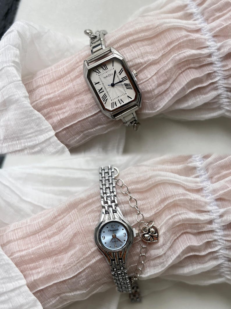 K&ouml;nnte beinhalten: Zwei silberne Armbanduhren mit Metallb&auml;ndern. Die obere Uhr hat ein wei&szlig;es Zifferblatt mit r&ouml;mischen Ziffern f&uuml;r die Stunden und den Text "NAIDU". Die untere Uhr hat ein blaues Zifferblatt mit arabischen Ziffern f&uuml;r die Stunden und den Text "Champagne".