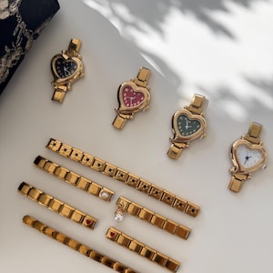 May include: Gold-toned heart-shaped watches with various colored faces, including black, red, green, and white. Several gold link bracelets with decorative elements, such as stars, pearls, and heart-shaped charms, are also displayed. The watches and bracelets are arranged on a white surface.