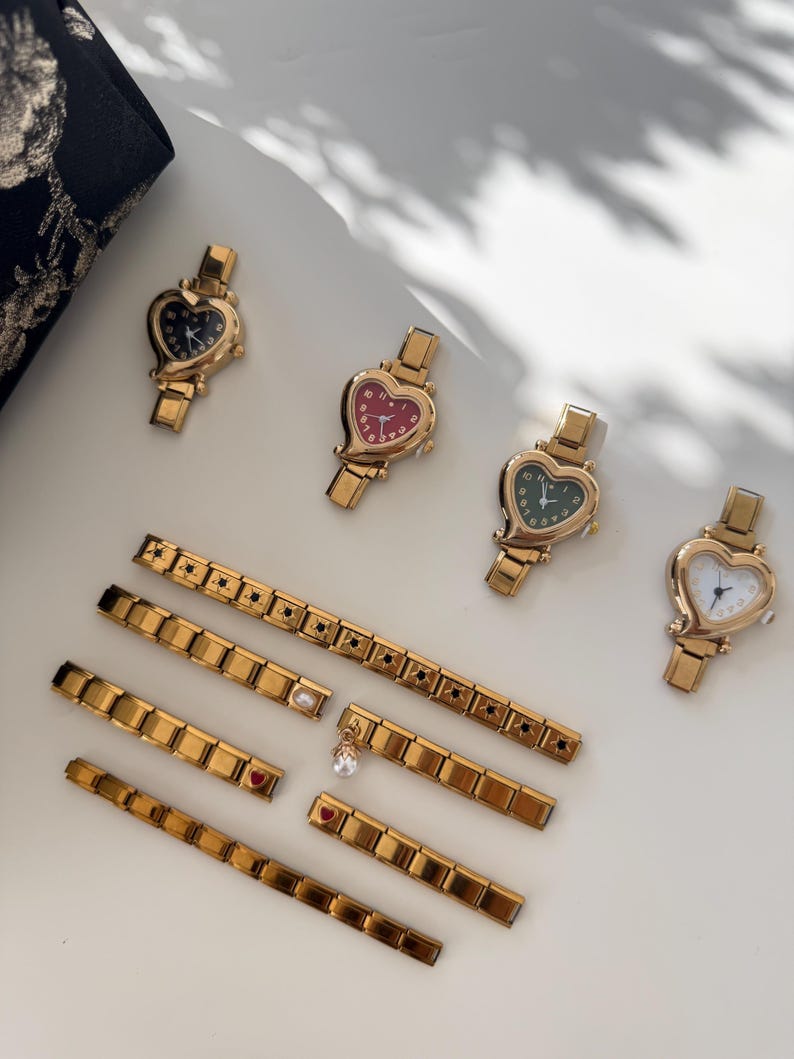 May include: Gold-toned heart-shaped watches with various colored faces, including black, red, green, and white. Several gold link bracelets with decorative elements, such as stars, pearls, and heart-shaped charms, are also displayed. The watches and bracelets are arranged on a white surface.