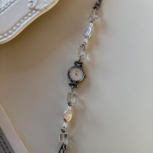 Pu&ograve; includere: Un orologio da polso argentato con quadrante bianco e cinturino con perline. Il cinturino presenta perline quadrate trasparenti, perle irregolari e piccole perline rotonde. L'orologio &egrave; su una superficie chiara.