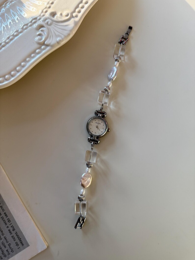 Pu&ograve; includere: Un orologio da polso argentato con quadrante bianco e cinturino con perline. Il cinturino presenta perline quadrate trasparenti, perle irregolari e piccole perline rotonde. L'orologio &egrave; su una superficie chiara.