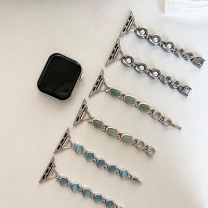 May include: A silver Apple Watch and three decorative watch bands. Two bands have clear, round stones, one has green oval stones, and two have blue oval stones. The bands are laid out on a white surface.