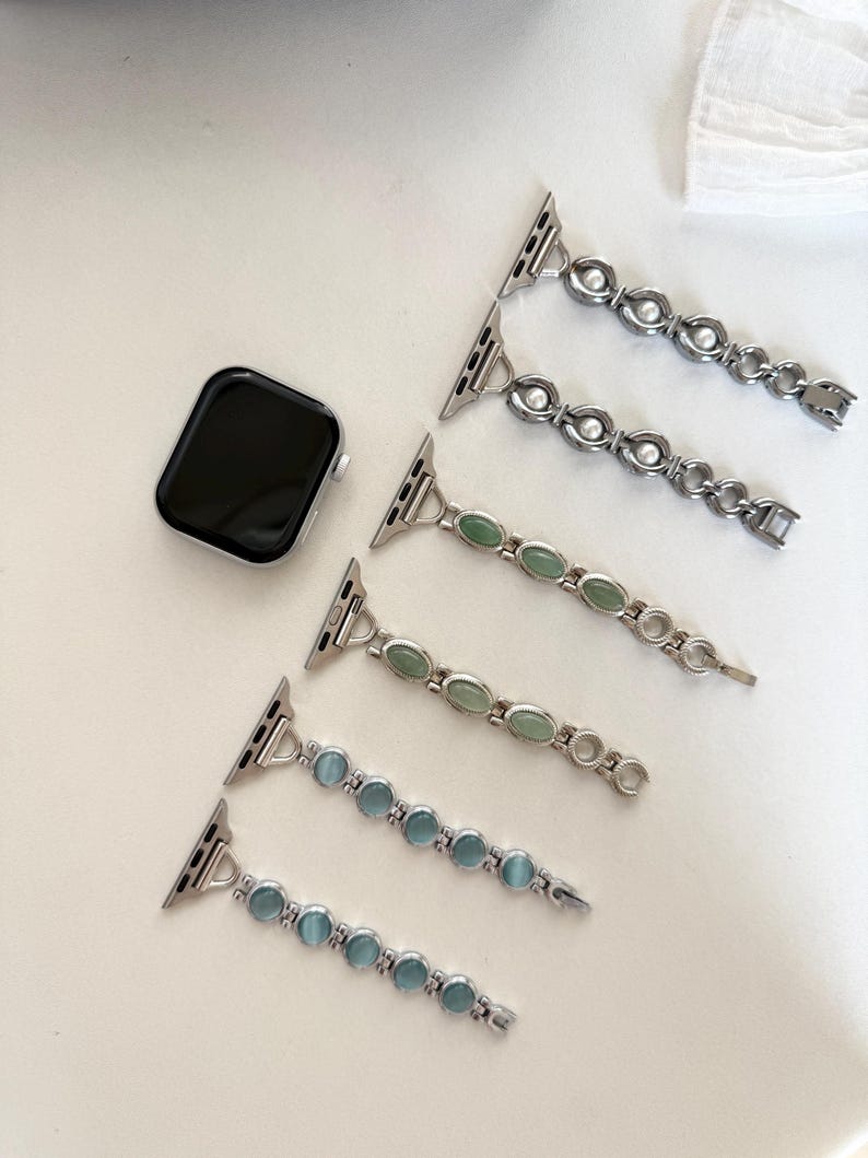 May include: A silver Apple Watch and three decorative watch bands. Two bands have clear, round stones, one has green oval stones, and two have blue oval stones. The bands are laid out on a white surface.
