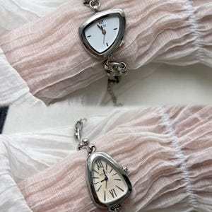 K&ouml;nnte beinhalten: Eine silberne Uhr mit einem dreieckigen Zifferblatt und r&ouml;mischen Ziffern. Die Uhr hat ein silbernes Kettenarmband.