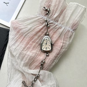 K&ouml;nnte beinhalten: Eine silberne Uhr mit einem einzigartigen, tropfenf&ouml;rmigen Zifferblatt und einem zarten Gliederarmband. Das Zifferblatt der Uhr hat r&ouml;mische Ziffern und einen silberfarbenen Stunden- und Minutenzeiger.