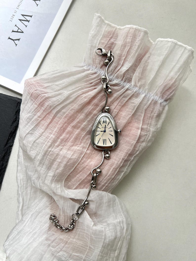 K&ouml;nnte beinhalten: Eine silberne Uhr mit einem einzigartigen, tropfenf&ouml;rmigen Zifferblatt und einem zarten Gliederarmband. Das Zifferblatt der Uhr hat r&ouml;mische Ziffern und einen silberfarbenen Stunden- und Minutenzeiger.