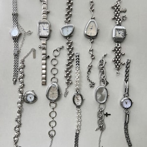 K&ouml;nnte beinhalten: Eine Sammlung von zehn silbernen Armbanduhren mit verschiedenen Band- und Zifferblattstilen. Die Uhren haben verschiedene Charms und Verzierungen, darunter Sterne, Herzen und Muscheln.