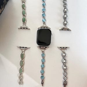 May include: An Apple Watch with several decorative bands. The bands are silver-toned with various gemstone and pearl accents. Gemstones include green and light blue oval-shaped stones. The watch face is black.