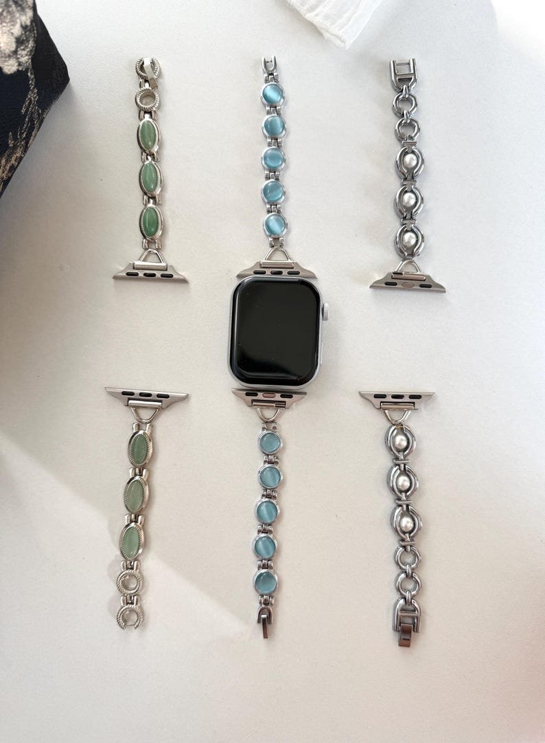 May include: An Apple Watch with several decorative bands. The bands are silver-toned with various gemstone and pearl accents. Gemstones include green and light blue oval-shaped stones. The watch face is black.