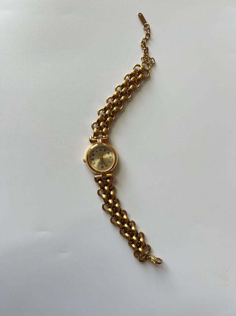 Puede incluir: Un reloj de pulsera dorado con una esfera redonda y una pulsera de eslabones.