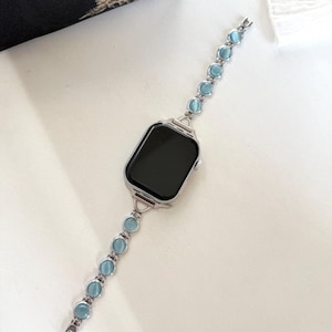 May include: Silver smartwatch with a black screen and a light blue beaded band. The band features round, light blue beads connected by silver links. The watch is displayed on a white surface.