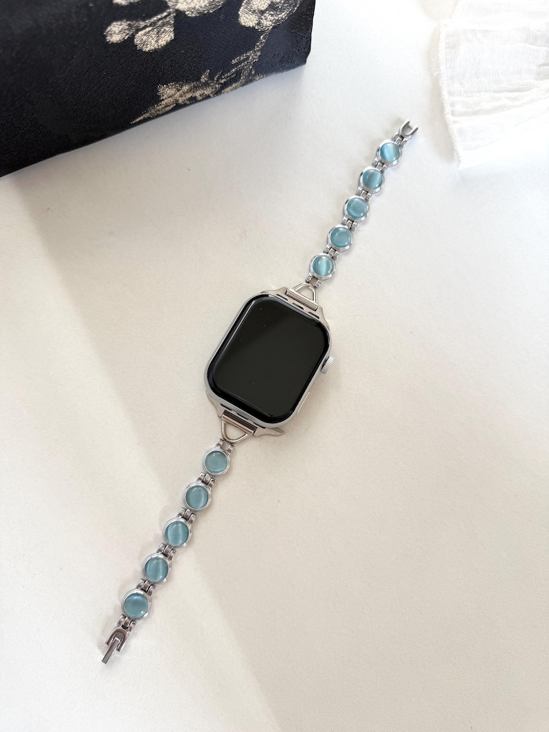 May include: Silver smartwatch with a black screen and a light blue beaded band. The band features round, light blue beads connected by silver links. The watch is displayed on a white surface.