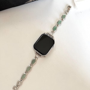 May include: Silver-toned watch band with oval, light green stones. The band is connected to a black-screened watch face. The watch band is on a white surface, with a black floral fabric in the background.