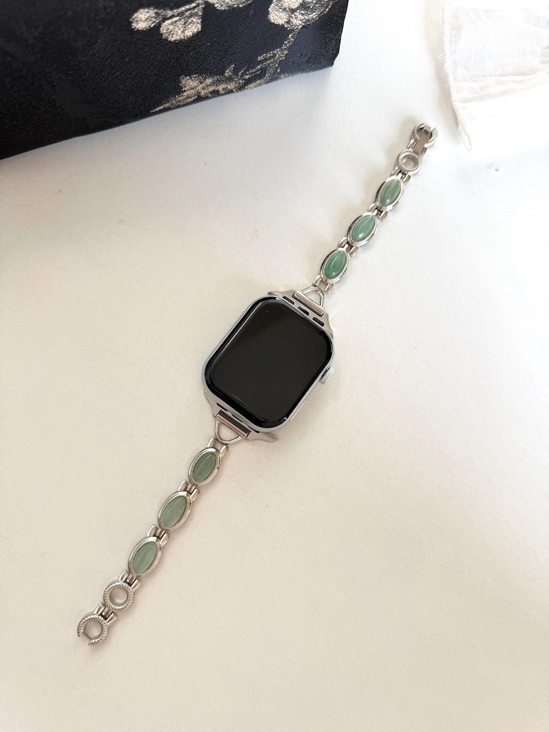 May include: Silver-toned watch band with oval, light green stones. The band is connected to a black-screened watch face. The watch band is on a white surface, with a black floral fabric in the background.