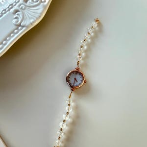 Pu&ograve; includere: Un delicato orologio da polso in oro rosa con quadrante bianco e lancette rosse. Il cinturino &egrave; composto da perline trasparenti e sferiche con dettagli dorati. Un accessorio alla moda.