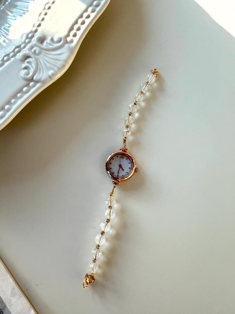 Pu&ograve; includere: Un delicato orologio da polso in oro rosa con quadrante bianco e lancette rosse. Il cinturino &egrave; composto da perline trasparenti e sferiche con dettagli dorati. Un accessorio alla moda.