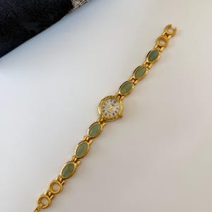 May include: A gold-toned wristwatch with a round face and a band featuring oval-shaped green stones. The watch face has Roman numerals and gold hands. The band is made of gold-colored metal with a clasp closure.