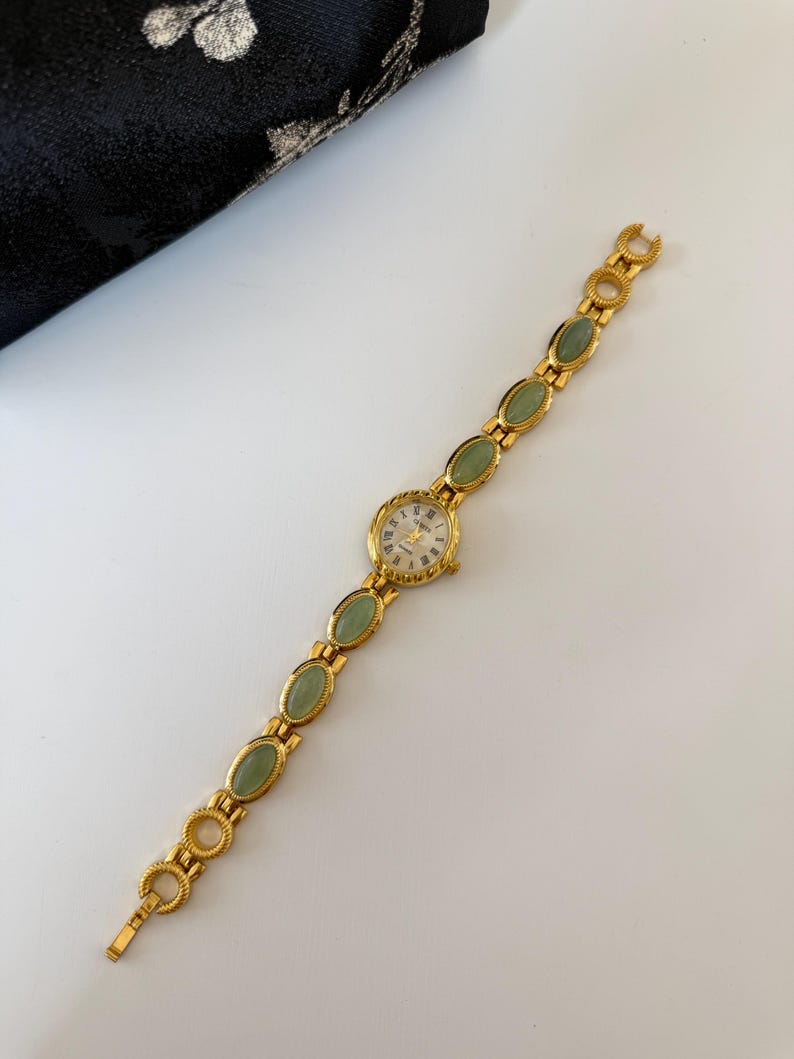May include: A gold-toned wristwatch with a round face and a band featuring oval-shaped green stones. The watch face has Roman numerals and gold hands. The band is made of gold-colored metal with a clasp closure.