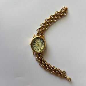 Puede incluir: Un reloj de pulsera dorado con una esfera redonda y marcadores de hora con n&uacute;meros romanos. El reloj tiene una pulsera de eslabones de cadena dorada.
