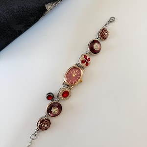 May include: A vintage-style wristwatch with a red face and gold-tone case. The watch band features decorative red and silver floral and jewel accents. The watch is a delicate piece of jewelry.