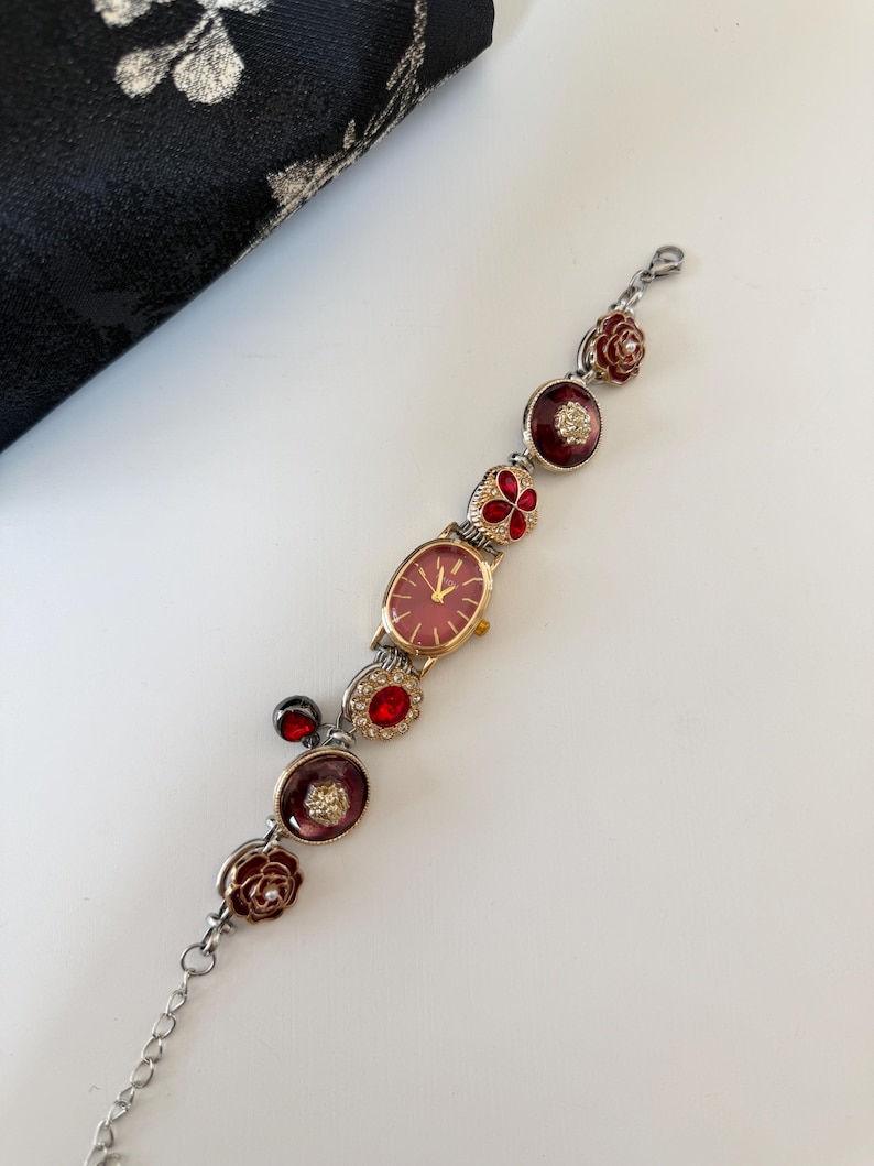 May include: A vintage-style wristwatch with a red face and gold-tone case. The watch band features decorative red and silver floral and jewel accents. The watch is a delicate piece of jewelry.