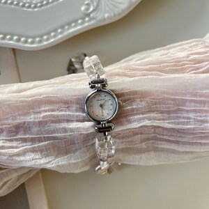 Pu&ograve; includere: Un orologio da polso argentato con quadrante rotondo e bianco. Il cinturino dell'orologio presenta dettagli chiari, rettangolari, simili a cristalli. L'orologio &egrave; esposto su un tessuto rosa chiaro e trasparente.