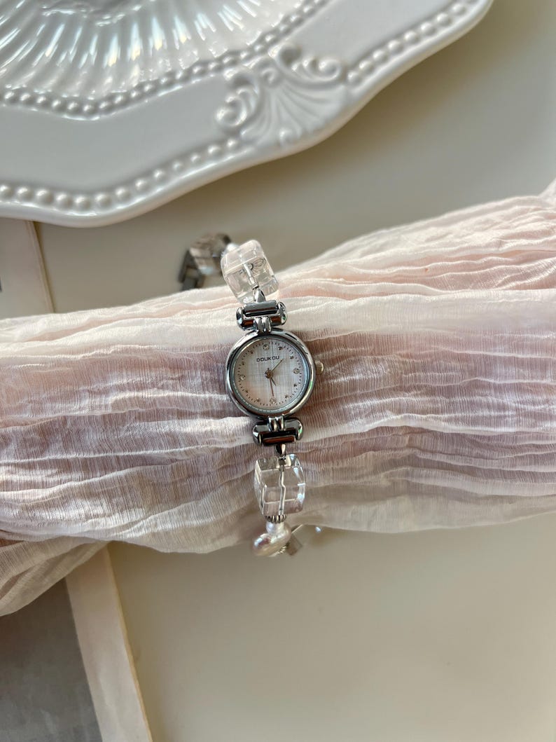 Pu&ograve; includere: Un orologio da polso argentato con quadrante rotondo e bianco. Il cinturino dell'orologio presenta dettagli chiari, rettangolari, simili a cristalli. L'orologio &egrave; esposto su un tessuto rosa chiaro e trasparente.