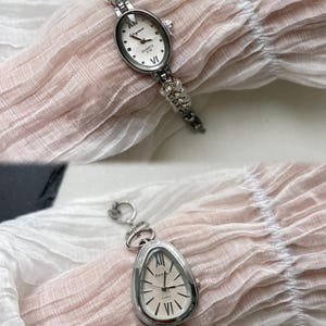 K&ouml;nnte beinhalten: Zwei silberne Uhren mit wei&szlig;en Zifferbl&auml;ttern und r&ouml;mischen Ziffern als Stundenmarkierungen. Die Uhr oben hat ein rundes Zifferblatt und ein Kettenarmband mit kleinen wei&szlig;en Perlen. Die Uhr unten hat ein ovales Zifferblatt und ein Kettenarmband mit gr&ouml;&szlig;eren silbernen Gliedern.