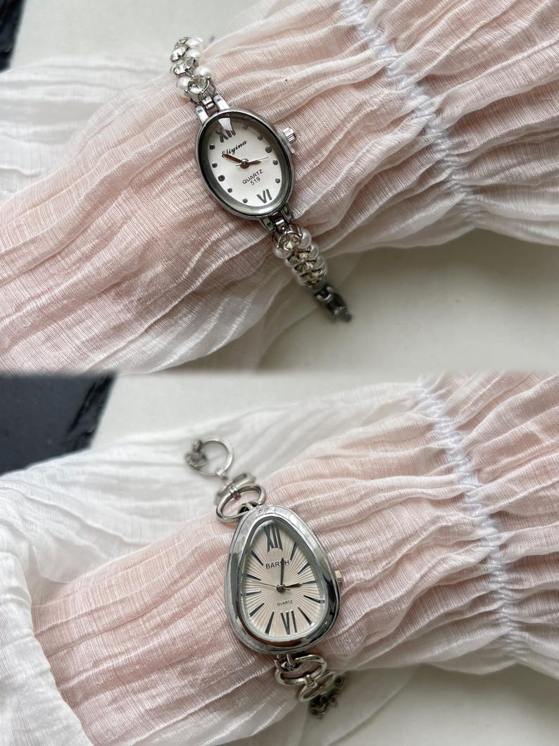 K&ouml;nnte beinhalten: Zwei silberne Uhren mit wei&szlig;en Zifferbl&auml;ttern und r&ouml;mischen Ziffern als Stundenmarkierungen. Die Uhr oben hat ein rundes Zifferblatt und ein Kettenarmband mit kleinen wei&szlig;en Perlen. Die Uhr unten hat ein ovales Zifferblatt und ein Kettenarmband mit gr&ouml;&szlig;eren silbernen Gliedern.