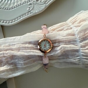 Pu&ograve; includere: Un orologio in oro rosa con quadrante rotondo e chiaro. Il cinturino dell'orologio &egrave; composto da perline di pietre preziose rosa e viola. L'orologio &egrave; esposto su un tessuto rosa chiaro.
