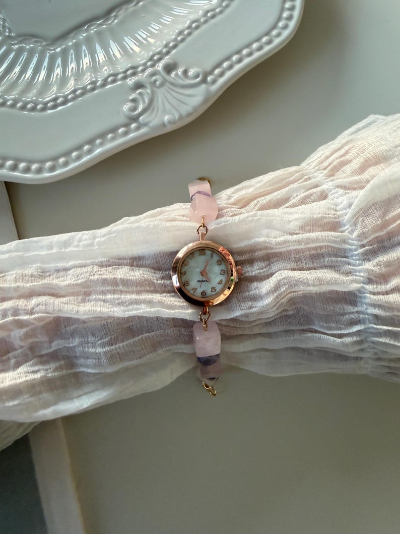 Pu&ograve; includere: Un orologio in oro rosa con quadrante rotondo e chiaro. Il cinturino dell'orologio &egrave; composto da perline di pietre preziose rosa e viola. L'orologio &egrave; esposto su un tessuto rosa chiaro.