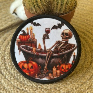 May include: Round Halloween patch with a black border. The patch features a skeleton relaxing in a bathtub filled with red liquid, holding a glass of wine. Pumpkins, candles, and bats complete the spooky scene.