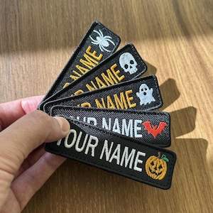 May include: Five rectangular black embroidered patches arranged in a fan shape. Each patch features the text YOUR NAME in white or gold thread, accompanied by a small Halloween-themed icon: a white spider, a white skull, a white ghost, a red bat, or an orange pumpkin.