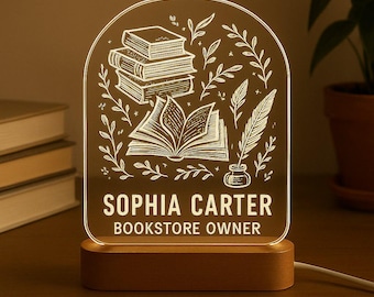 May include: A clear acrylic LED lamp with a wooden base. The lamp features an engraved design of books, leaves, and a quill. The text "SOPHIA CARTER BOOKSTORE OWNER" is displayed below the design. The lamp is plugged into an electrical outlet.