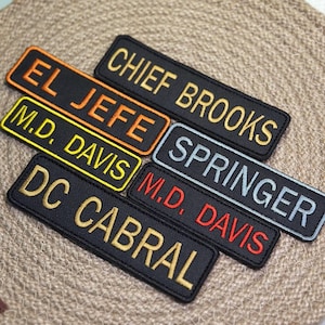Pode incluir: Uma coleção de patches retangulares pretos com texto bordado em várias cores. Os patches exibem nomes e títulos como "CHIEF BROOKS", "EL JEFE" e "M.D. DAVIS". Os patches estão dispostos em uma superfície texturizada de tons neutros.
