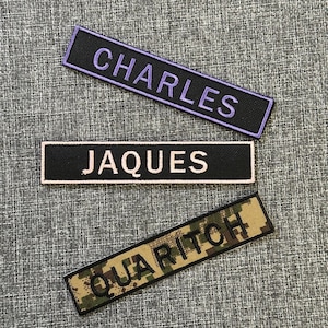 Pode incluir: Tr&ecirc;s patches retangulares bordados com nomes. O patch superior tem "CHARLES" em fio roxo sobre fundo preto. O patch do meio tem "JAQUES" em branco sobre preto. O patch inferior mostra "QUARITCH" em verde sobre um padr&atilde;o de camuflagem.
