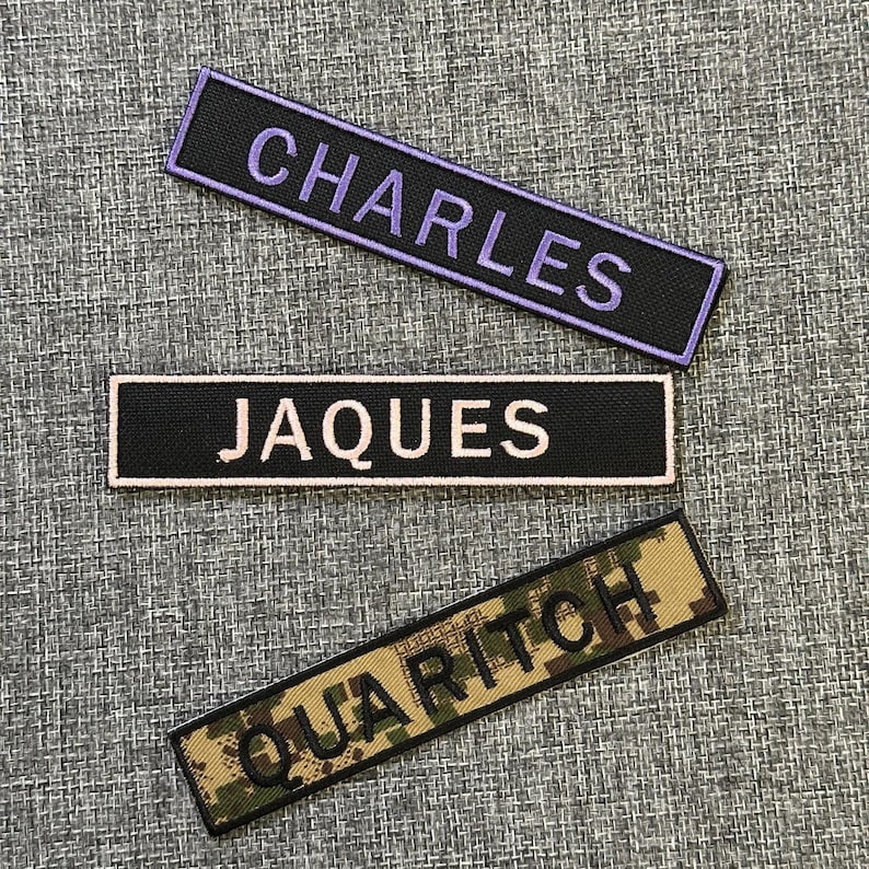 Pode incluir: Tr&ecirc;s patches retangulares bordados com nomes. O patch superior tem "CHARLES" em fio roxo sobre fundo preto. O patch do meio tem "JAQUES" em branco sobre preto. O patch inferior mostra "QUARITCH" em verde sobre um padr&atilde;o de camuflagem.