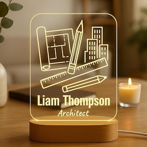 Puede incluir: Lámpara de escritorio de arquitecto personalizada con base de madera. El panel acrílico transparente presenta un diseño grabado de planos, lápices y edificios. Se muestra el nombre "Liam Thompson" y el título "Architect".