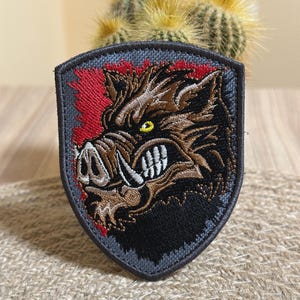 Può includere: Toppa ricamata a forma di scudo, raffigurante una testa di cinghiale dettagliata. Il cinghiale è marrone con zanne bianche e occhi gialli, su sfondo rosso e nero. La toppa ha un bordo grigio.