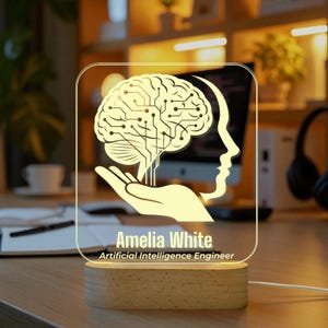May include: A lit acrylic desk lamp with a brain design and circuit board pattern, held by a hand, and a profile of a face. The lamp sits on a wooden base and has the name "Amelia White" and title "Artificial Intelligence Engineer".