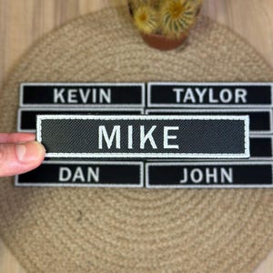 Könnte beinhalten: Schwarz-weiße Namensschilder mit weißem Text. Der Name "MIKE" wird von einer Hand gehalten. Weitere Namen sind "KEVIN", "TAYLOR", "DAN" und "JOHN". Die Aufnäher befinden sich auf einer gewebten Oberfläche.