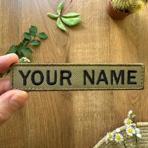 Può includere: Una toppa rettangolare verde oliva con la scritta "YOUR NAME" ricamata in nero. La toppa è tenuta in mano su uno sfondo di legno, con rametti di verde e piccoli fiori bianchi.