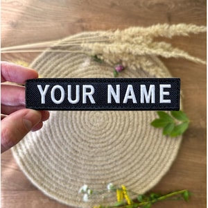Pode incluir: Um patch retangular preto com o texto "YOUR NAME" em letras brancas. O patch &eacute; segurado em uma m&atilde;o. O fundo &eacute; neutro com elementos decorativos.