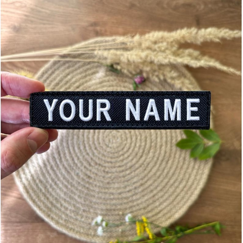 Pode incluir: Um patch retangular preto com o texto "YOUR NAME" em letras brancas. O patch &eacute; segurado em uma m&atilde;o. O fundo &eacute; neutro com elementos decorativos.