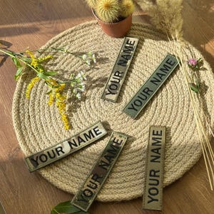 May include: Five rectangular patches with the text "YOUR NAME" in black lettering. The patches have a camouflage pattern in shades of green, brown, and tan. They are arranged on a round, woven placemat. A small potted cactus and dried flowers are also present.