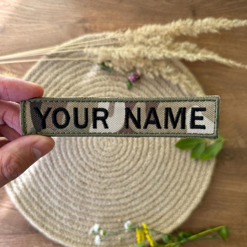 Pode incluir: Um patch retangular com o texto "YOUR NAME" em preto, bordado em um padr&atilde;o de camuflagem. O patch &eacute; segurado em uma m&atilde;o, com um fundo neutro de um tapete tecido e plantas secas.