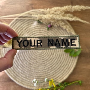 Pode incluir: Um patch retangular com o texto "YOUR NAME" em preto, bordado em um padr&atilde;o de camuflagem. O patch &eacute; exibido em um fundo neutro, com gram&iacute;neas secas e uma esteira tecida.